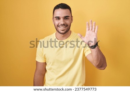 Young hispanic man standing over yellow background waiving saying hello happy and smiling, friendly welcome gesture 