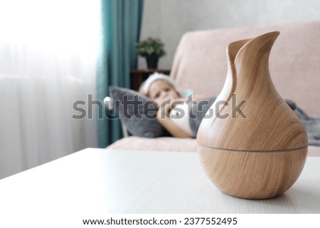 A diffuser in the form of a wooden jug on a light table. Little girl in the background. The concept of air humidification in the house, aromatherapy for stress and depression, healthy sleep. Royalty-Free Stock Photo #2377552495