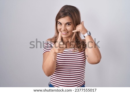Young hispanic woman standing over isolated background smiling doing talking on the telephone gesture and pointing to you. call me. 