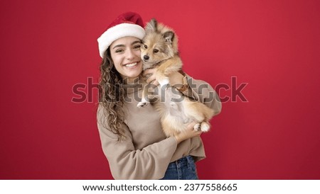 Young hispanic woman with dog smiling confident wearing christmas hat over isolated red background