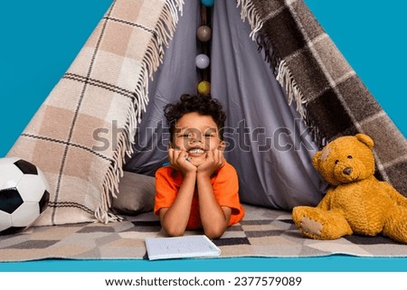 Photo of boy lying in homemade tepee writing in copybook isolated blue color background