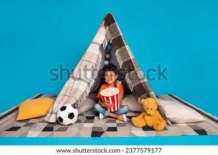 Photo of sweet adorable boy watching television eating pop corn in handmade tent isolated blue color background