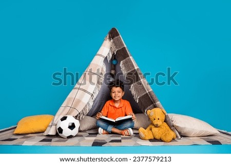 Photo of cute sweet boy playing in diy tent reading fairytale isolated blue color background