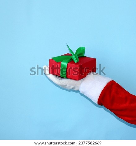 Santa Claus's hand holds a red gift box on a blue background. Gift present for Happy New Year concept. Close-up.