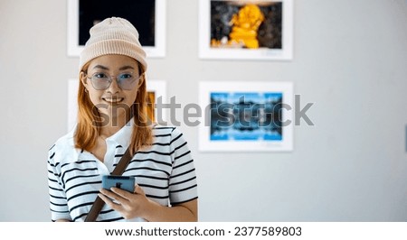 Visitor woman smiling at picture art gallery collection in front framed paintings pictures she holding mobile phone, Asian female watching at photo frame with smartphone at artwork gallery show
