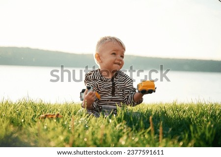Against lake behind. Cute little boy is having fun outdoors on the ground with green grass.