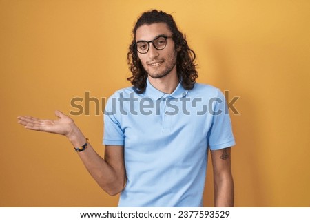 Young hispanic man standing over yellow background smiling cheerful presenting and pointing with palm of hand looking at the camera. 
