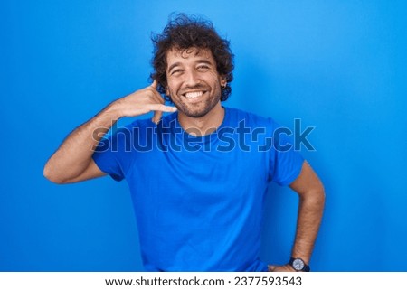 Hispanic young man standing over blue background smiling doing phone gesture with hand and fingers like talking on the telephone. communicating concepts. 