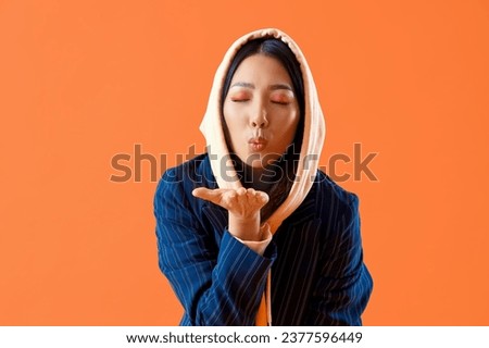 Young Asian woman in hoodie blowing kiss on orange background
