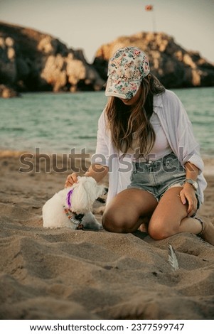 Photo of woman in hat sitting with her sweet white Maltese dog on the beach
