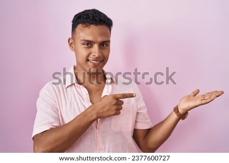Young hispanic man standing over pink background amazed and smiling to the camera while presenting with hand and pointing with finger. 