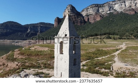 A close-up photo of the Sant Romà de Sau church, showing only its bell tower. The bell tower has become visible due to this year's drought, with green mountains and cliffs in the background.