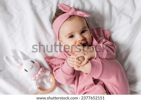 Baby in a pink jumpsuit with an eco-friendly wooden rattle is lying in bed