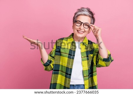 Photo of optimistic nice woman wear checkered shirt touch glasses palm demonstrate offer empty space isolated on pink color background