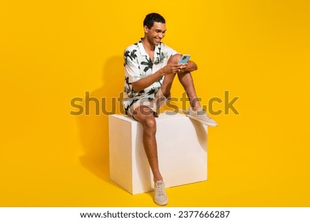 Full size photo of handsome young guy sit white podium hold device wear trendy palms print outfit isolated on yellow color background