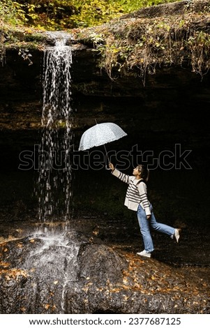 Picture on an autumn day of a young lady with a white umbrella in her hand, who is next to a waterfall in nature.