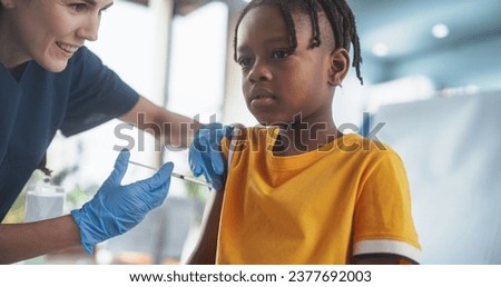 Young African American Boy Sitting In The Chair In Bright Hospital And Getting Polio Vaccine. Caucasian Female Nurse Performing Injection. Professional Woman Talks To Worried Kid, Calms Him Down. Royalty-Free Stock Photo #2377692003