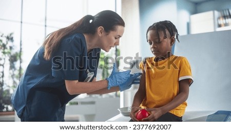 Young African American Boy Sitting In The Chair In Bright Hospital And Getting Polio Vaccine. Caucasian Female Nurse Is Performing Injection. Professional Woman Talks To Worried Kid, Calms Him Down. Royalty-Free Stock Photo #2377692007