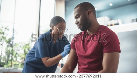 Happy African Man Sitting In The Chair In Bright Hospital And Getting His Hepatitis B Vaccine. Professional Black Female Nurse Is Performing Injection And Putting Patch On. Public Healthcare Concept Royalty-Free Stock Photo #2377692011