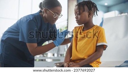 Young African American Boy Sitting In The Chair In Bright Hospital And Getting His Polio Vaccine. Female Black Nurse Is Performing Injection. Professional Woman Talks To Worried Kid And Calms Him Down Royalty-Free Stock Photo #2377692027