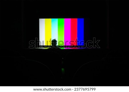 Silhouette of a man, a lone spectator sitting in a chair in the cinema hall against the background of a screen with an image of a color test