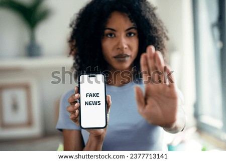 Shot of serious woman holding the mobile phone with the message fake news while doing stop sign with hand at home