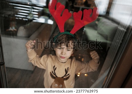 Child in knitted sweater and deer headband stands near window, looks outside. Realistic lifestyle photo.