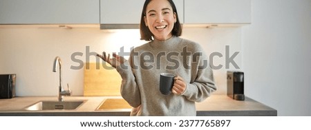 Portrait of stylish asian woman standing in kitchen with mug, drinking coffee, explaining smth, showing banner or advertisement.
