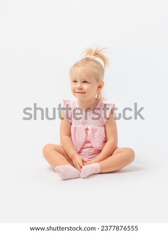 A cute blonde girl 1-2 years old in a pink dress is sitting on the floor smiling and looking away on a white background. Copy space.