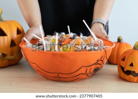 Woman holding a Halloween candy bowl filled with sweets for trick-or-treating. Female hands and Halloween-themed decorative treat bowl container. Jack-o'-lantern pumpkins placed around the table.