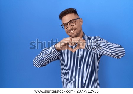 Hispanic man with beard wearing glasses smiling in love doing heart symbol shape with hands. romantic concept. 
