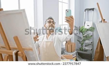Smiling young, grey-haired hispanic man artist captures a joyful selfie with smartphone amidst paintbrushes and canvas in art studio