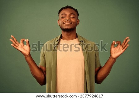 Portrait of peaceful african man enjoying calm meditation in studio with green background. Spiritual young guy holding hands in mudra gesture and keeping eyes closed.