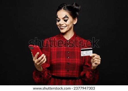 Young woman with Halloween makeup face art mask wear clown costume red dress hold use mobile cell phone credit card isolated on plain solid black background studio portrait Scary holiday party concept
