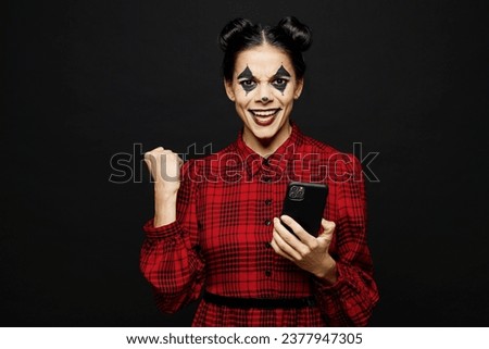 Young winner woman with Halloween makeup face art mask wears clown costume red dress hold use mobile cell phone isolated on plain solid black background studio portrait. Scary holiday party concept