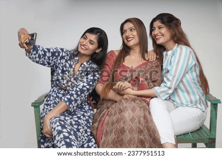 Indian beautiful happy women taking selfie picture on mobile phone while sitting on bench isolated on grey studio background.