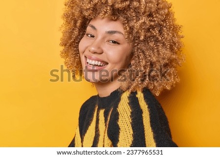 Sideways shot of cute cheerful European woman with curly bushy hair smiles broadly has white teeth expresses sincere emotions and feelings dressed in casual jumper isolated over yellow background Royalty-Free Stock Photo #2377965531