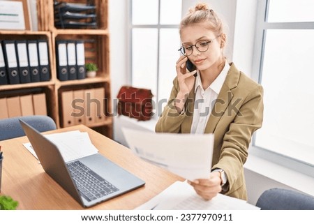 Young blonde woman business worker talking on smartphone reading document at office