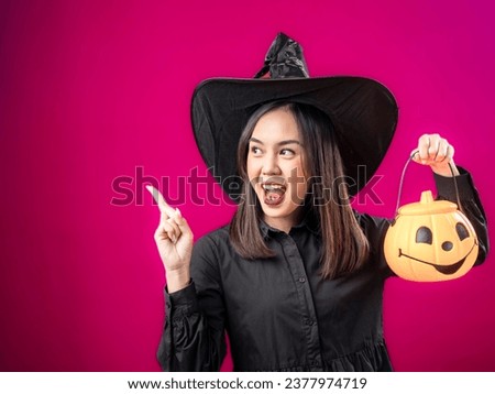 Portrait of an Asian Indonesian woman wearing a Halloween-themed costume with a witch hat, pointing towards something with her hand while holding a pumpkin. Isolated against a magenta background.