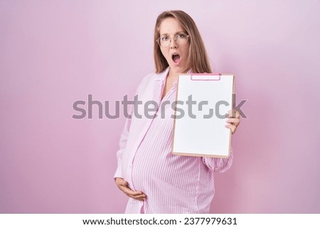 Young pregnant woman holding clipboard in shock face, looking skeptical and sarcastic, surprised with open mouth 
