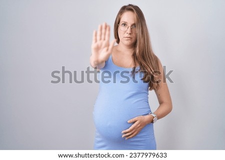 Young pregnant woman standing over white background doing stop sing with palm of the hand. warning expression with negative and serious gesture on the face. 