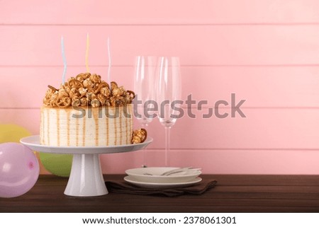 Caramel drip cake decorated with popcorn and pretzels near balloons and tableware on wooden table, space for text