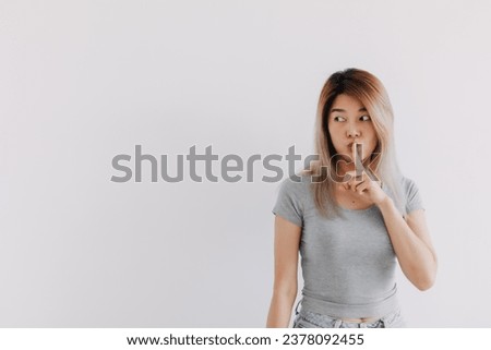 Asian Thai woman wear grey, hold fingers on lips, showing shh, hush quiet sign, making gesture silence, keep secret, looking at empty space, isolated on white background wall.