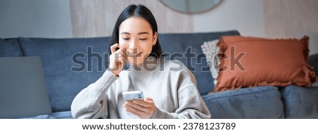 Portrait of smiling asian woman looking intrigued at smartphone screen, interested with smth on mobile phone, sitting coy on floor in living room. Royalty-Free Stock Photo #2378123789