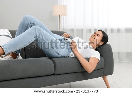 Young woman struggling to squeeze into tight jeans while lying on sofa at home Royalty-Free Stock Photo #2378137773