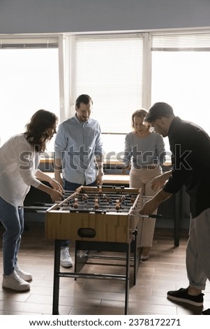 Four office employees, friends, workmates engaged in foosball game, two colleagues cheer on teammates enjoy lunch break, having fun together during workday, vertical shot. Hobby, friendship at work Royalty-Free Stock Photo #2378172271