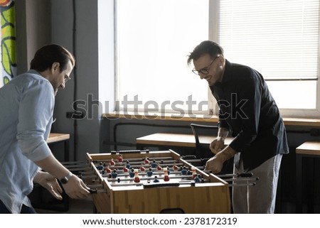 Two positive excited colleagues having fun at workplace during lunch break, millennial friends enjoying break at work, entertaining in office, playing table football, competing in board soccer game Royalty-Free Stock Photo #2378172319