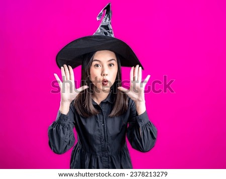 Portrait of an Asian Indonesian woman wearing a Halloween-themed costume with a witch hat, blowing a kiss with her hand near her head. Isolated against a magenta background.