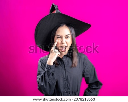 Portrait of an Asian Indonesian woman wearing a Halloween-themed costume with a witch hat, striking a teasing pose by pointing and winking with one hand. Isolated against a magenta background.