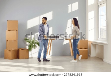 Happy young family couple moving into new home. Cheerful man and woman homeowners carrying table in empty living room with cardboard boxes. Moving day, real estate concept Royalty-Free Stock Photo #2378241915